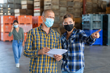 Man and young woman in face masks talking about documentation while standing in warehouse. Another woman talkin on phone in background.