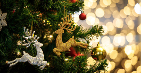 Close up shot of Golden and silver glossy Christmas eve decor reindeer jumping model hanging decorating on green Xmas pine tree branch with sphere balls and ice flakes on blurred bokeh background