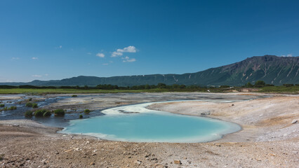 Hot natural springs flow into an amazing turquoise lake. Sulphurous deposits on the soil around. A picturesque mountain range against the blue sky. Kamchatka. The caldera of the extinct volcano Uzon