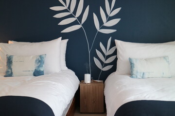 There are hand-painted leaves graffiti on the dark blue wall. There are two comfortable beds in front. The pillowcases are rendered and look very beautiful.