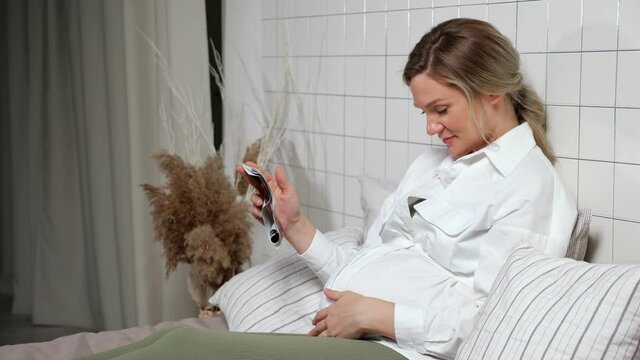 Blonde haired young pregnant woman in white shirt and green tights sits on bed and strokes tummy looking at ultrasound picture leaning on pillows