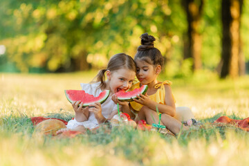 Two little mixed race kids consist of european and caucasian girls smiling with happiness, fun amusement, playing, sitting for picnic and eating piece of watermelon fruit in outdoor garden