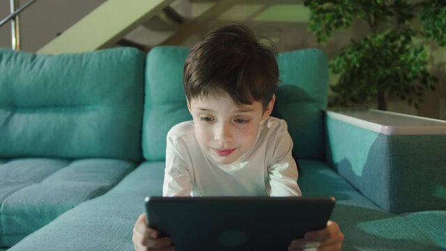 Curious Little Kid Boy Using Digital Pad Entertainment Device Lying On Sofa Alone. Small Boy Hold Pad Computer Playing Games At Home Watching Video. Children Tech Addiction Concept