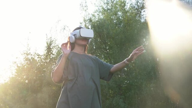 A girl in virtual reality glasses stands against the background of green trees. He moves his hands through the air, trying to catch a sunbeam. The concept of the future. High quality 4k footage