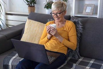 An older woman with short blonde hair sits on sofa in living room and chats with friend on laptop over video chat