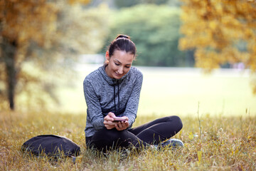 Young woman with happy face dressed in outdoor sportswear sitting on the grass in park and checks messages on smartphone