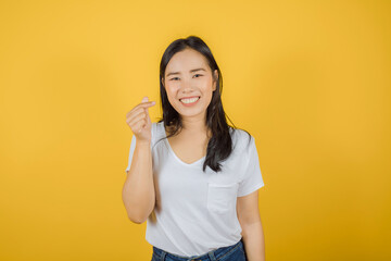 Portrait of young beautiful Asian woman with long dark hair wears white t-shirt showing mini heart gesture isolated on yellow background