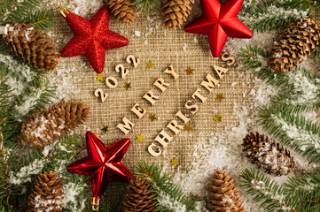 Merry Christmas and New Year 2022. festive background with red stars, snow-covered branches of spruce and cones on burlap. top view.