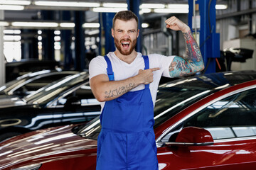 Strong young technician car mechanic man in denim overalls white t-shirt show point on biceps...