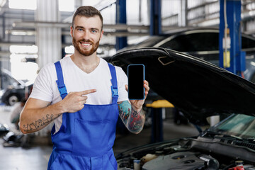 Young technician car mechanic man in overalls white t-shirt use hold point finger on mobile cell...
