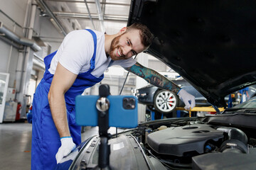 Young troubleshooter technician car mechanic man in blue overalls white t-shirt talk mobile cell on phone stand show process fix problem with raised hood work in vehicle repair shop workshop indoors
