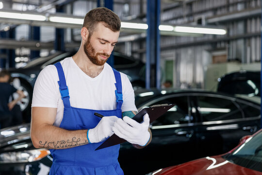 Young Serious Car Mechanic Man In Denim Blue Overalls White T-shirt Gloves Estimate Hold Clipboard With Papers Document Work In Light Modern Vehicle Repair Shop Workshop Indoors. Tattoo Translate Fun.