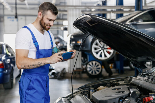 Young Professional Technician Car Mechanic Man In Blue Overalls White T-shirt Take Photo Picture On Mobile Cell Phone Fix Problem With Raised Hood Bonnet Work In Vehicle Repair Shop Workshop Indoor