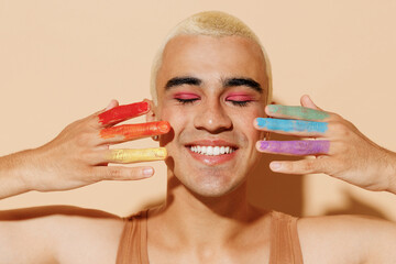 Young smiling happy blond latin gay man with pink eyeshadow make up fingers painted in rainbow flag...