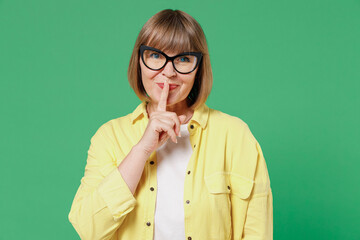 Elderly secret caucasian woman 50s wearing glasses yellow shirt says hush be quiet with finger on...
