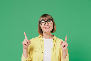 Elderly smiling fun happy woman 50s in glasses yellow shirt point index finger overhead on workspace area copy space mock up isolated on plain green color background studio People lifestyle concept.