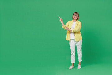 Full body elderly smiling happy cool woman 50s in glasses yellow shirt point index finger aside on workspace area mock up isolated on plain green background studio portrait People lifestyle concept.