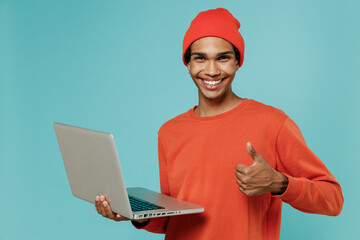 Young smiling happy african american man in orange shirt hat hold use work laptop pc computer...