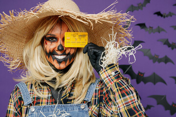 Close up fun young woman with Halloween makeup mask in straw hat scarecrow costume cover eye with credit bank card isolated on plain dark purple background studio Celebration holiday party concept
