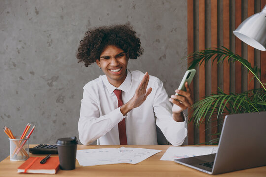 Young displeased stressed african american employee business man in classic shirt tie talk speak on mobile cell phone do stop palm gesture refusing sit work at white office desk with pc laptop inside.