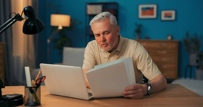 Shot of elderly businessman sitting in front of laptop with documents. Man working remotely in evening. Elderly man works in home office at computer conducts conferences via internet on video camera