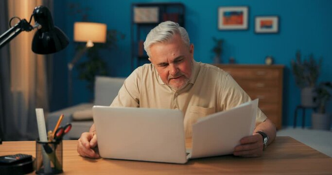 An elderly businessman reads company documents. Man laughing, happy with company's achievements. Smiling boss reading employee reports talk to employees through webcam, internet connection