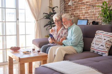 Smiling senior couple sitting on sofa at home using phone. White-haired elderly enjoying technology and retirement. Bright light from window, brick wall