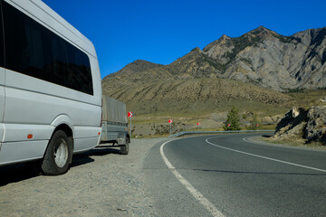 Obraz premium a white minibus with a trailer is standing on the side of the highway among the mountains. The road turns around a bend. Against the background of steep high mountains and clear blue sky