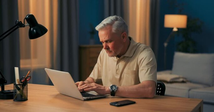 Elderly Man With Gray Hair Works On Laptop At Desk And Lamp In Evening. Mature Businessman Happy To Work Online, Remotely From Home. A Men Running An Online Business. Looking At The Camera