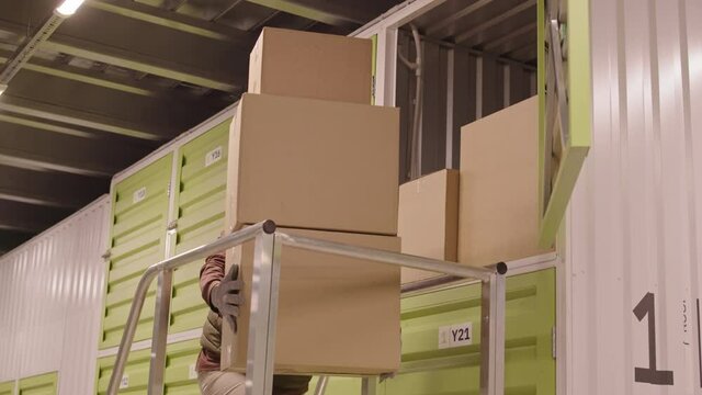 Locked-down of young Caucasian female warehouse worker holding stack of cardboard boxes, going up on ladder, then dropping them