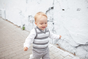 Fototapeta premium Stylish male toddler dressed fashion clothes, grey jeans, sweater and walking against the street wall