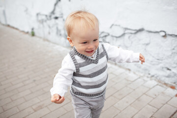 Stylish male toddler dressed fashion clothes, grey jeans, sweater and walking against the street wall