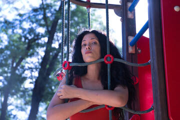 Middle-aged adult Hispanic woman with black curly hair, hugging the ropes of a climbing apparatus, shyly. Concept games, shyness, looks, sensations.