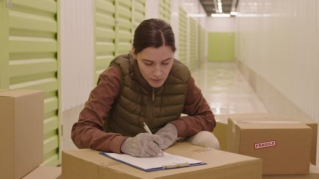 Medium long of young Caucasian woman surrounded by cardboard boxes in storage hallway, writing list on paper on clipboard