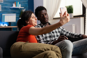Cheerful interracial couple taking pictures with smartphone at home. Multi ethnic adults creating memories and selfies with modern gadget in hand and technology. Mixed race happy partners