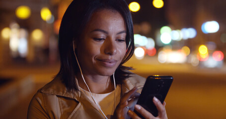 Bokeh shot of mixed-race woman in earphones using smartphone outdoors o in night city
