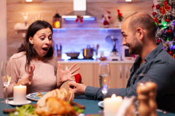 Happy cheerful man proposing girlfriend to marry him using diamond luxury engagement ring celebrating christmas holiday spending winter season together. Joyful couple sitting at xmas dinning table