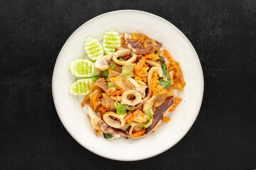 Pad See Ew Pla Muek, Thai food, stir fried soy sauce noodle with squid, egg, lettuce, carrot served with cucumber on dark tone texture background, top view