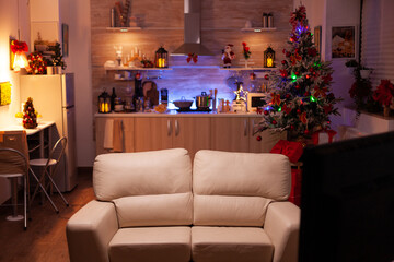 Empty xmas living room with nobody in it ready for winter holiday at home. In background x-mas decorated kitchen with modern furniture. Christmas ornaments at home. December festive decorations