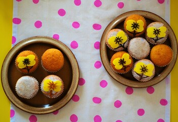 Plates of muffins decorated with powdered sugar and yellow icing in the shape of yellow flowers on a white tablecloth with pink polka dots on a yellow table