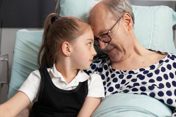 Caring cheerful granddaughter touching forehead with sick elderly grandfather supporting him during...