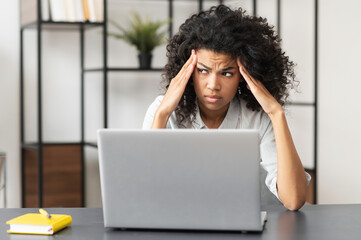 Tired African-American businesswoman in casual shirt sitting at the desk in front of laptop, upset...