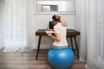 Woman sits on a fitness ball in headphones and works at home during quarantine