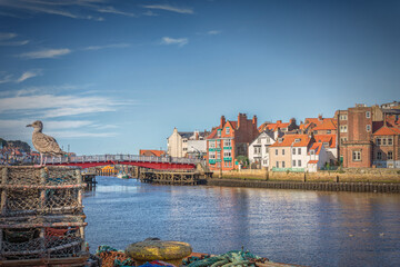Buildings line a quay and a red bridge spans the harbour. Lobster pots and a seagull are in the foreground and a sky with light cloud is above.