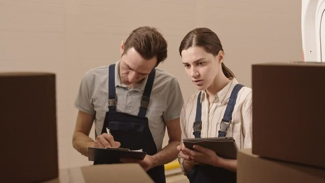 Medium view from inside back of vehicle of Caucasian woman and man wearing worker overalls checking packages, taking notes, talking before delivery