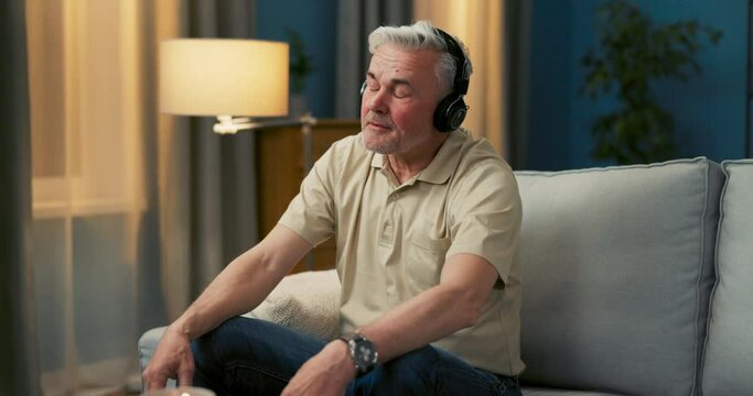 An elderly man is sitting on a couch in a comfortable position, wearing wireless headphones, listening to favorite songs, singing, nodding to the rhythm of the music, enjoying the track