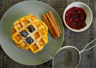 Belgian gluten-free pumpkin waffle, sprinkled with powdered sugar and blueberries, lies on a gray plate next to a strainer and jam on a wooden table, top view. vegan breakfast cooked at home