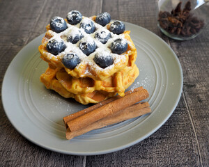 Belgian gluten-free pumpkin waffle, sprinkled with powdered sugar and blueberries, lies on a gray plate on a wooden table, side view. vegan breakfast cooked at home