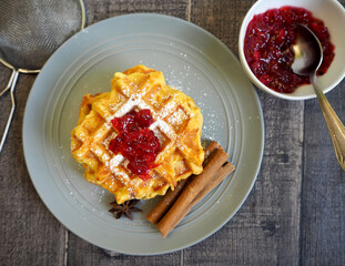 on a gray plate are Belgian gluten-free pumpkin waffles, sprinkled with powdered sugar and red currant jam, with cinnamon sticks. top view