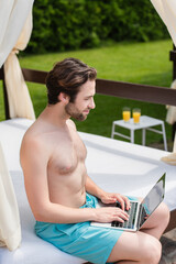 Side view of man in swimming trunks using laptop on lounge bed outdoors